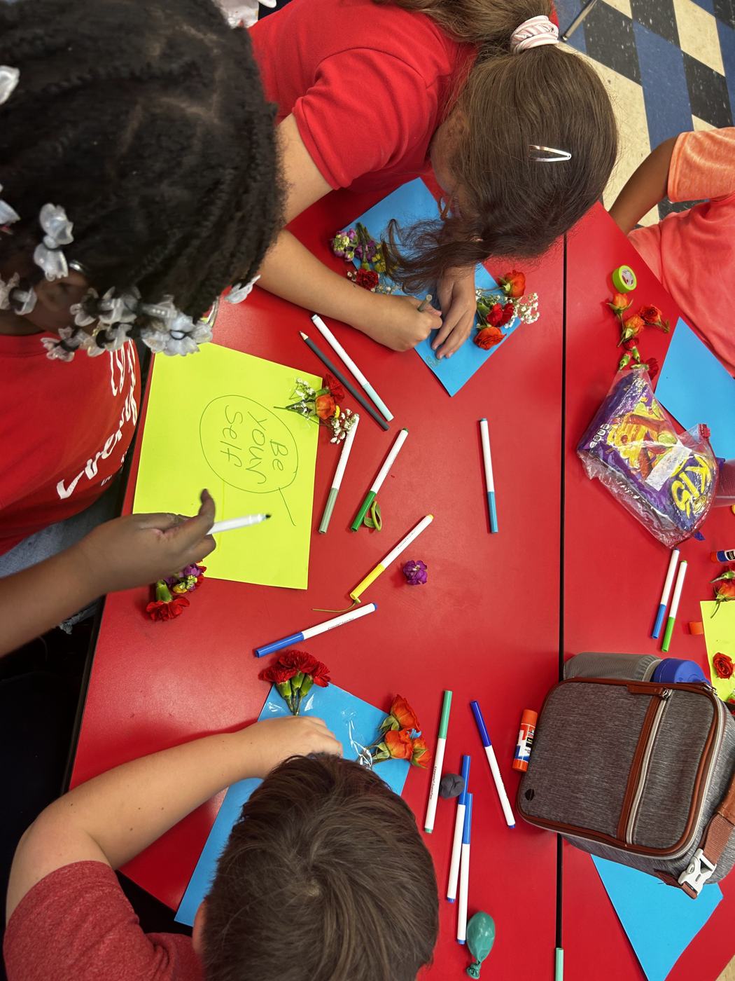 Students drawing at a red table covered in markers and flowers